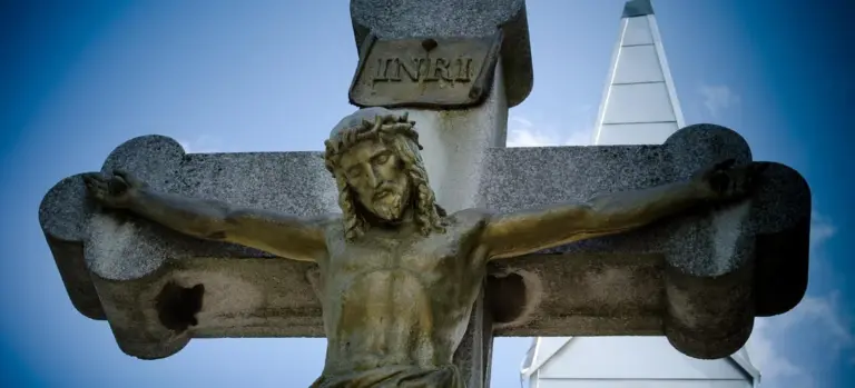 Outdoor crucifix statue with a cross and a church steeple in the background