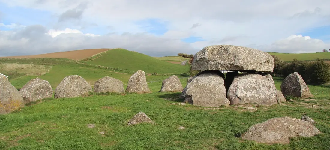 A field of ancient megalithic dolmens with a prominent capstone resting on vertical stones, set in green grass under a partly cloudy sky.