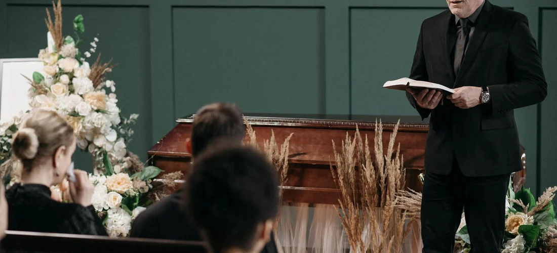 Funeral service scene: a speaker in a black suit reads from a book at a lectern, while attendees sit facing the front, with pale floral arrangements in the background.