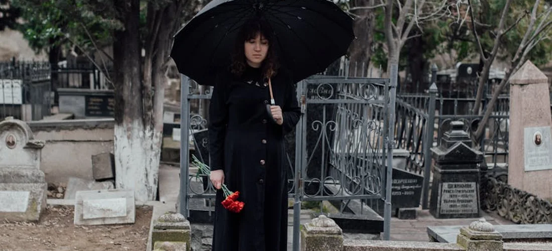 A person dressed in a dark coat stands in a cemetery holding a red bouquet under a large black umbrella, with tombstones and bare trees in the background.