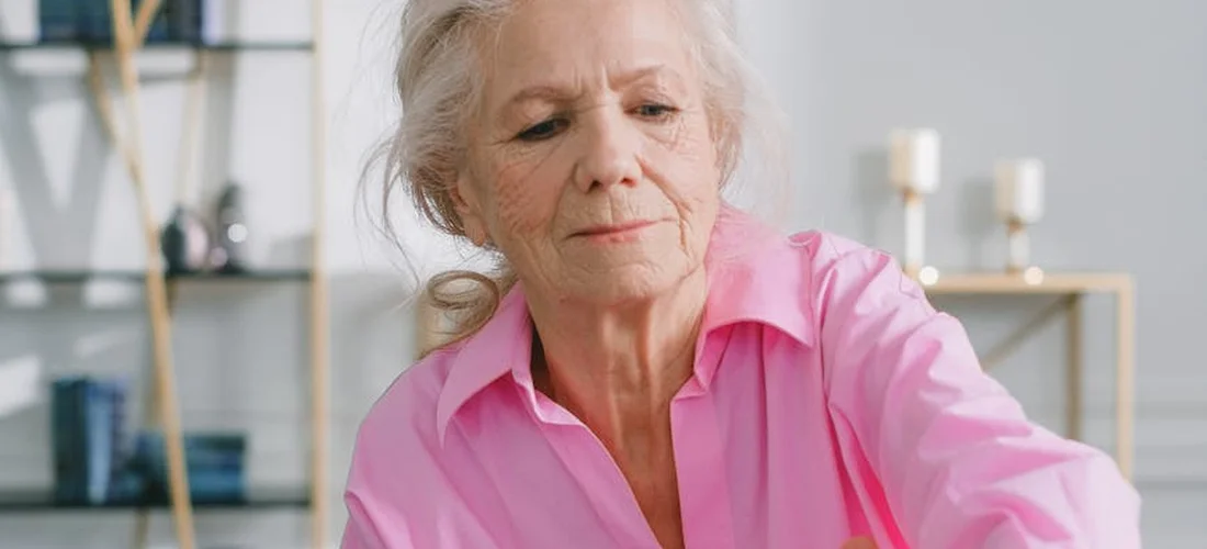 An elderly woman wearing a pink blouse sits indoors, looking thoughtful and contemplative.