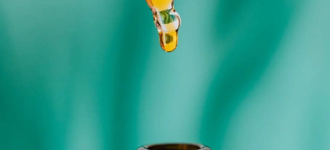 Close-up of a glass dropper releasing yellow liquid toward an open container against a teal background.