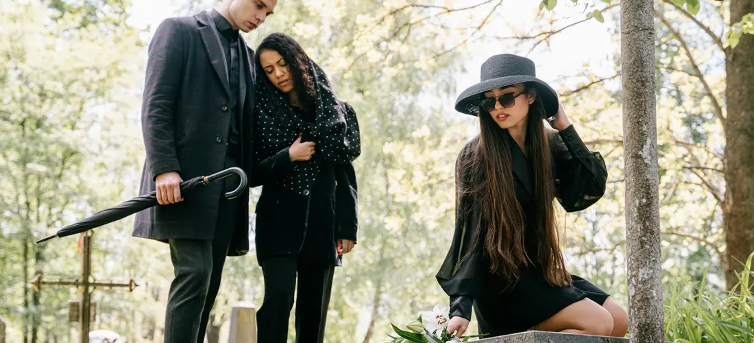Three people in a cemetery-like outdoor setting: a man in a suit stands with a shovel, while two women in dark clothing comfort each other, one sitting nearby with a bouquet.
