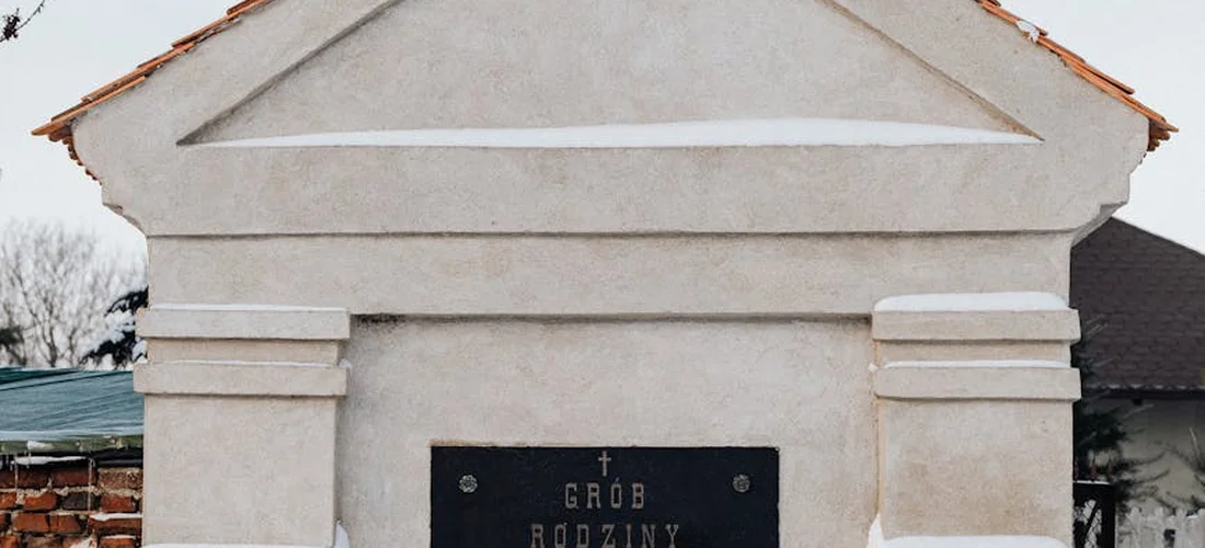 White family tomb in a cemetery with a dark plaque reading 'Grób Rodziny' (Family Tomb) in Polish.