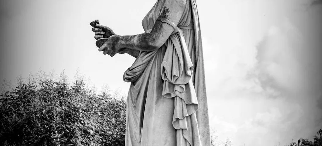 Black-and-white statue of a draped figure standing in a memorial garden, hands clasped near the waist.