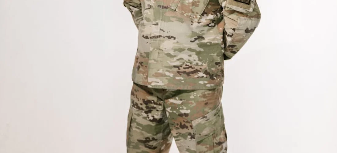 A service member in camouflage uniform stands with hands behind their back, symbolizing quiet respect during a military funeral.