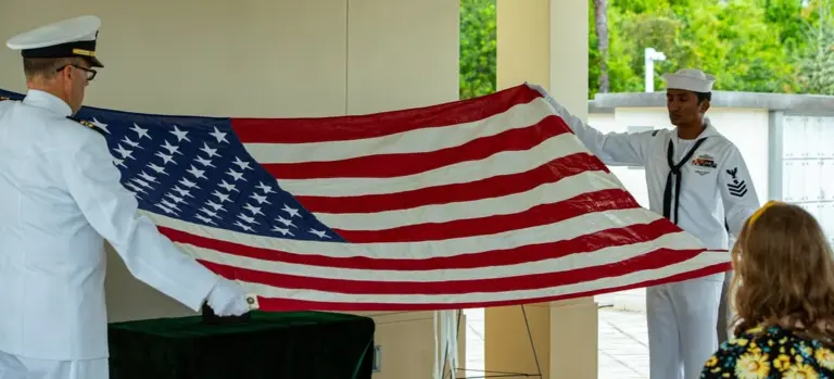 Two naval officers in white uniforms hold a large American flag during a flag-folding ceremony at a funeral or memorial service, with a guest seated nearby.