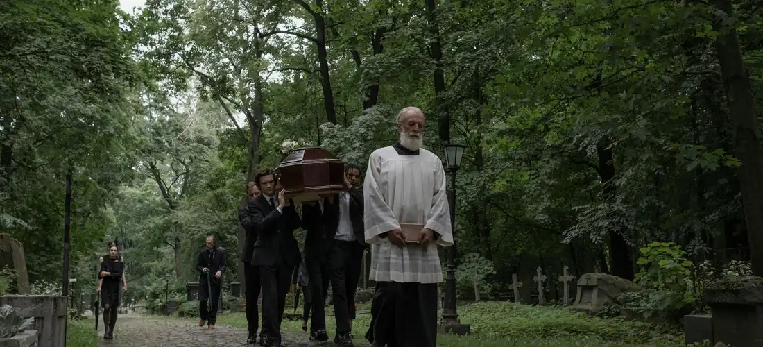 A funeral procession through a shaded forest cemetery with pallbearers carrying a coffin, led by a clergyman in a white robe.