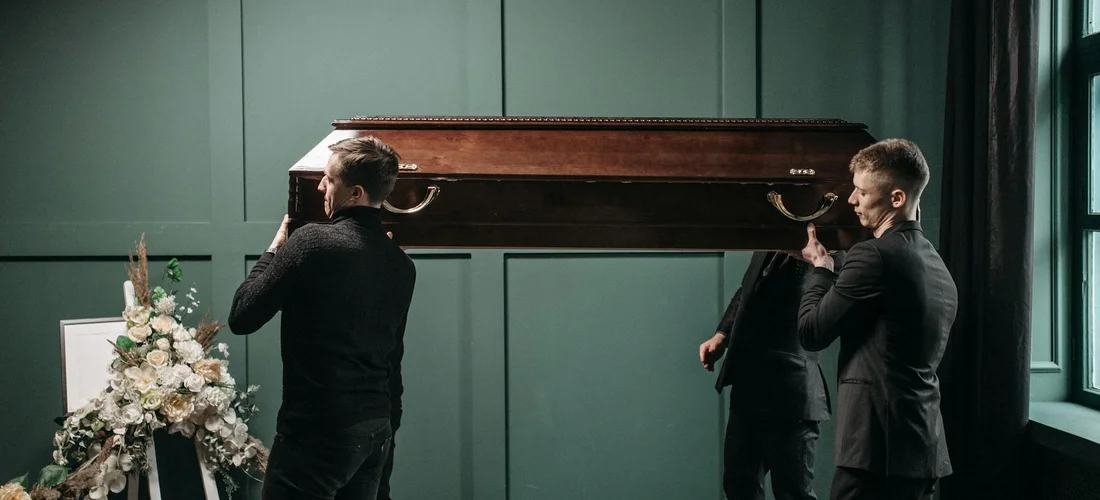 Two pallbearers in dark suits lift a wooden coffin in a green-walled room, with a floral arrangement nearby.