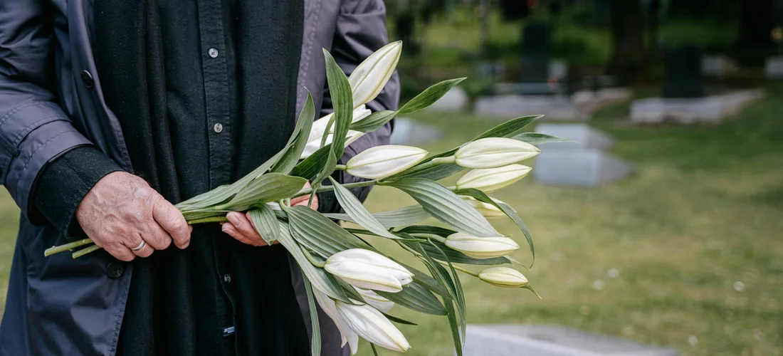 A person in dark clothing holds a bouquet of white lilies, with a cemetery in the blurred background.
