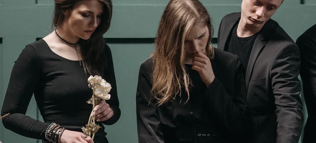 Three mourners dressed in dark, conservative clothing at a funeral, conveying somber attire and modest styles.