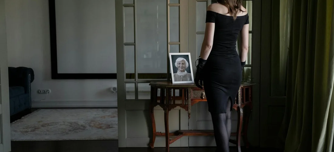 Woman in a black dress stands with her back to the camera in a living room, looking at a framed photo on a table.