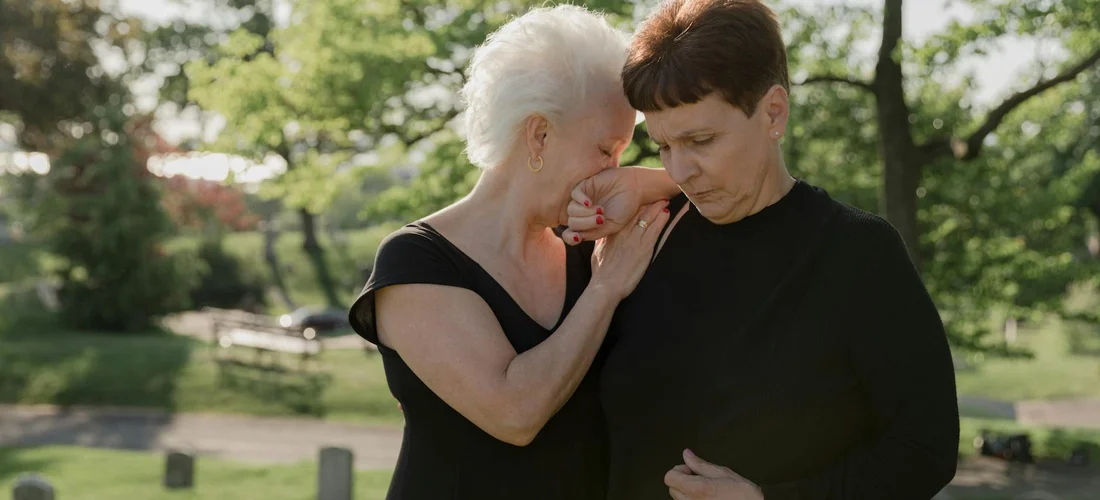 Two women dressed in black share a tender moment of comfort outdoors, symbolizing support during mourning.