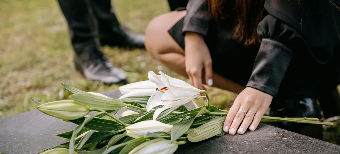 Close-up of white lilies laid on a dark memorial marker while hands in black sleeves rest nearby, a quiet funeral scene