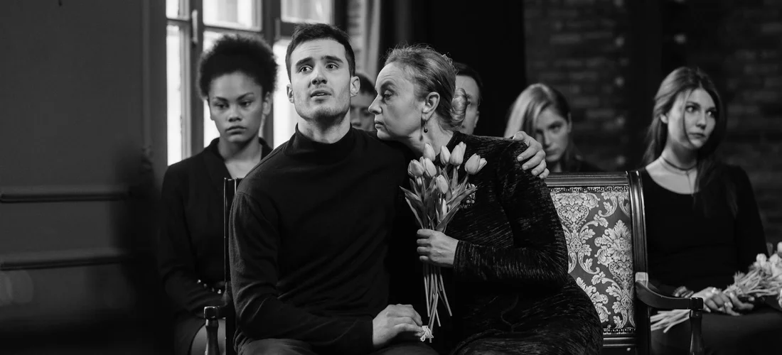 Black-and-white photo of a group of people in a funeral setting. A man sits in the foreground while a woman whispers to him, holding flowers; others sit nearby.