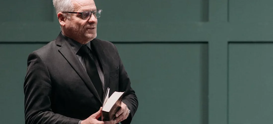 Man in a dark suit holding a book, standing against a muted green wall.