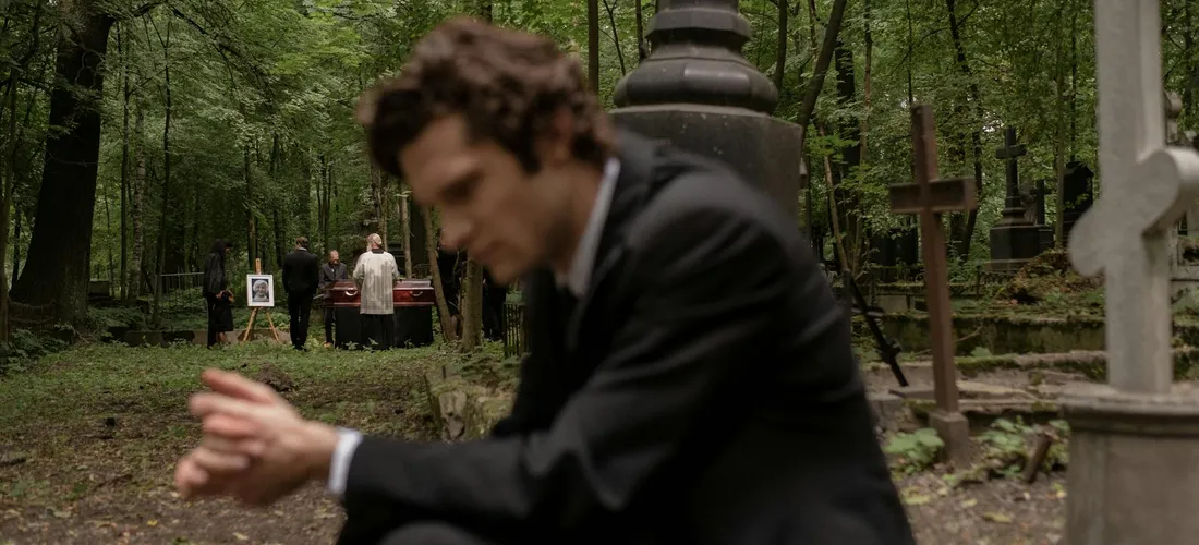 A man in a suit stands blurred in the foreground at a cemetery, with gravestones and greenery in the background and a small group of people visible in the distance.