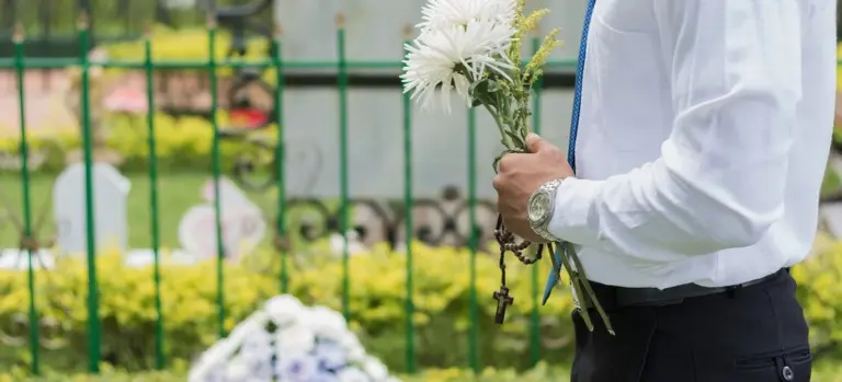 A person in a white shirt holds a bouquet of white flowers at a cemetery near a grave with a green fence in the background.