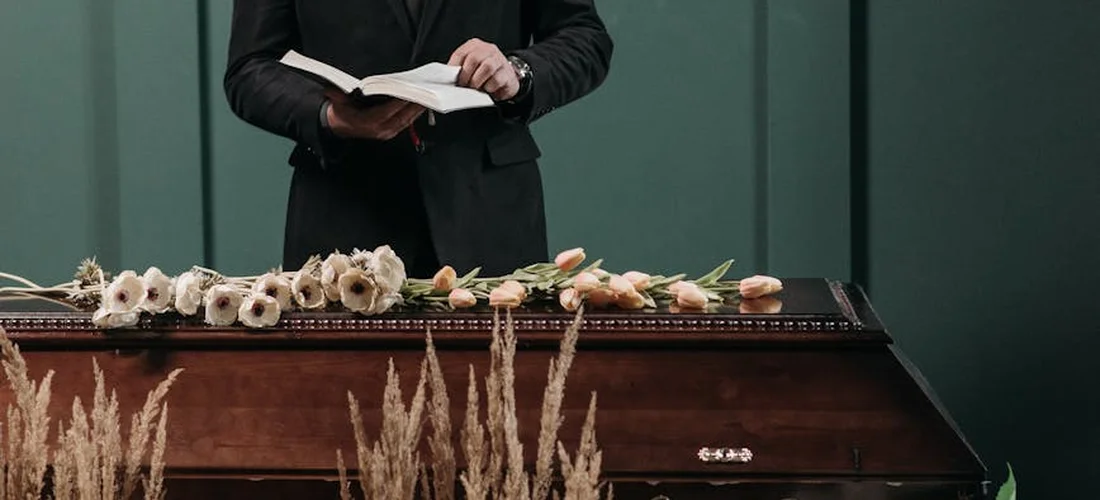 A professional in a dark suit stands beside an open book atop a decorated coffin with pale flowers.