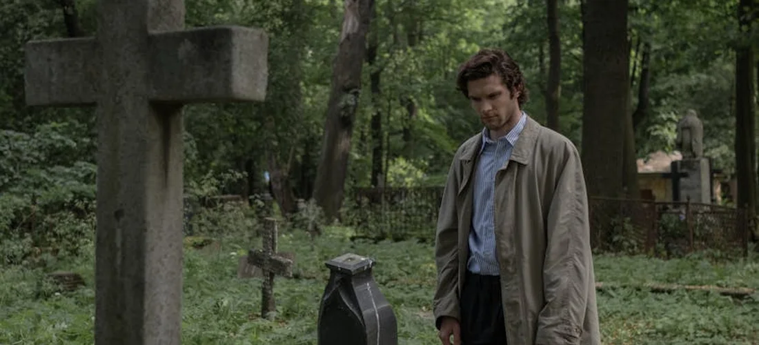 A man in a beige trench coat stands by a gravestone in a quiet cemetery, surrounded by trees.