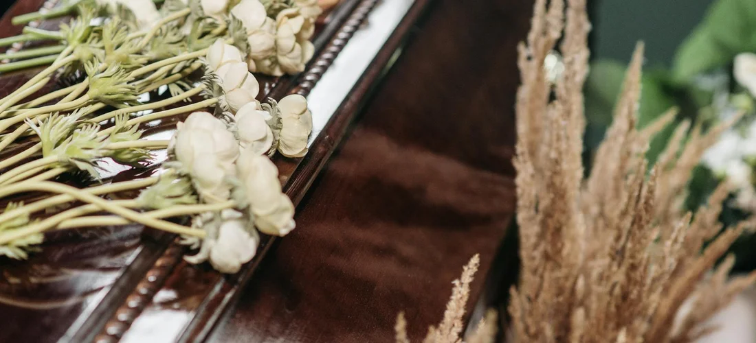 Close-up of white flowers and dried grasses resting on a polished wooden surface, suggesting a funeral setting.