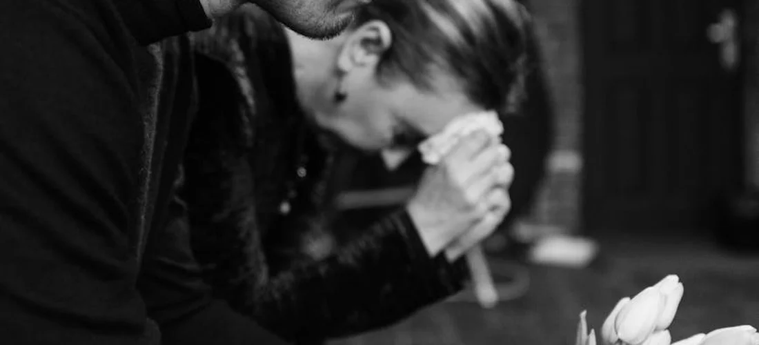 A grayscale photo of a grieving person sitting, hands clasped near their face, with a white flower in the foreground.