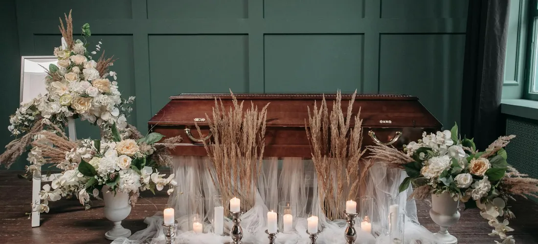 Funeral casket surrounded by white and cream floral arrangements and candles in a dark green room.