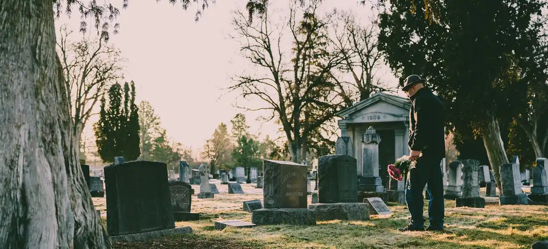 Person standing in a cemetery with a bouquet, surrounded by gravestones and trees on a sunny day.
