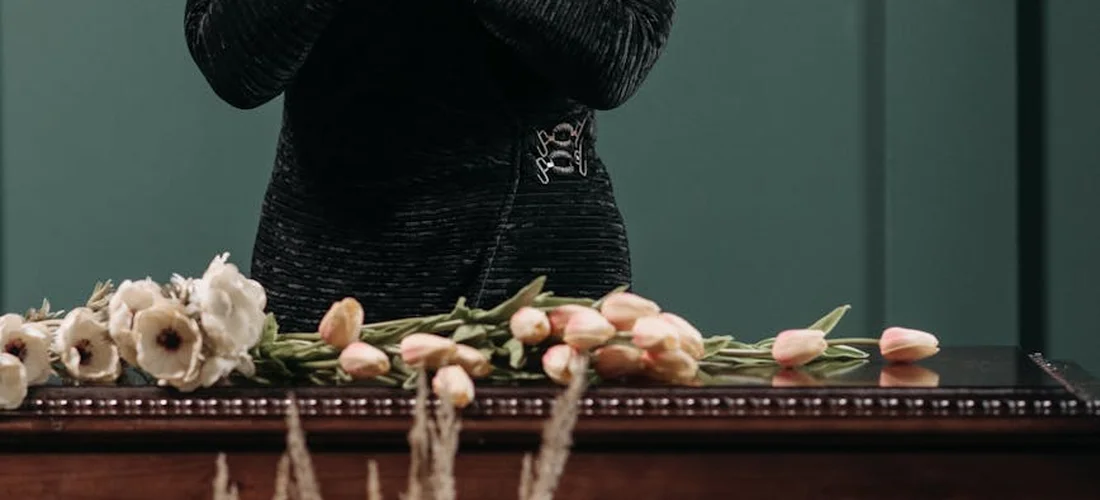 A long floral spray rests across a casket, featuring white blooms and pale pink tulips, with a person in dark clothing visible in the background.