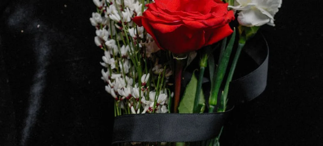 Bouquet featuring a red rose and white filler flowers wrapped with a black ribbon against a dark background.
