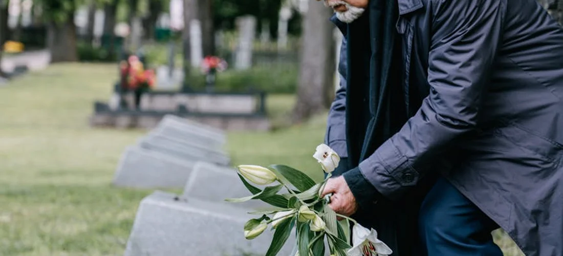 A person in a dark jacket bending toward a row of gravestones in a cemetery park, holding a bouquet of flowers.