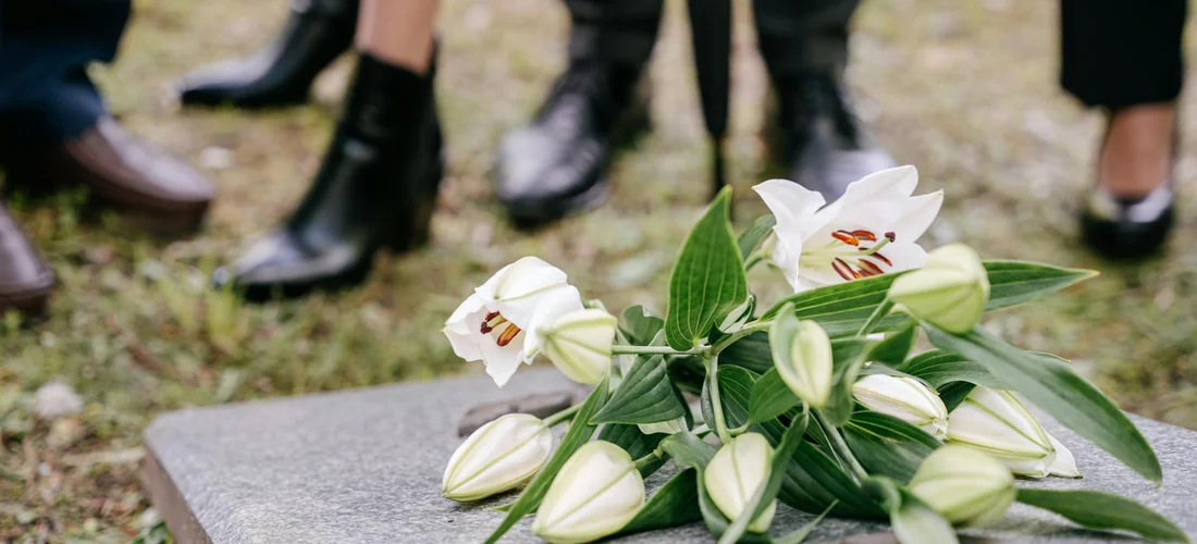 White lilies arranged on a gravestone in a cemetery, with mourners' feet visible in the background.