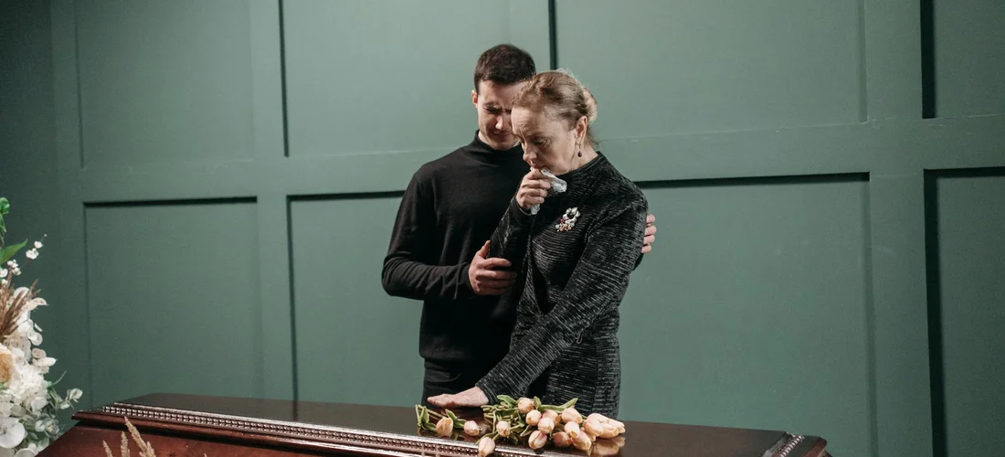 A man comforts a grieving woman beside a closed casket at a funeral home.