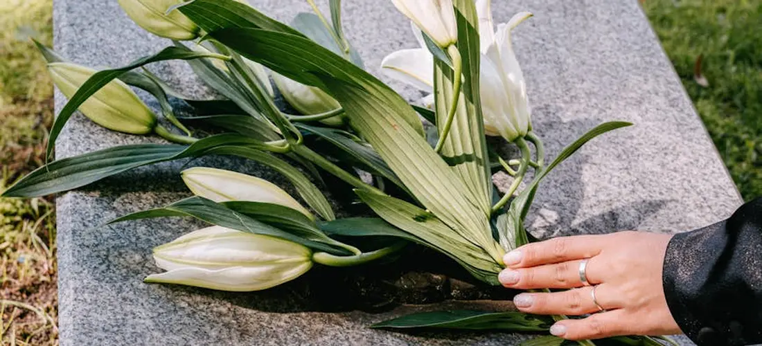 Close-up of a hand with a ring touching a gravestone with white lilies laid across the stone, symbolizing memorials and funeral planning.