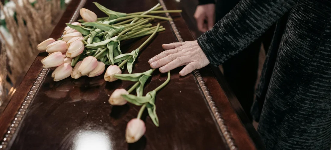 A hand rests on a dark coffin beside a bouquet of pale pink tulips, capturing a moment of reflection at a funeral.