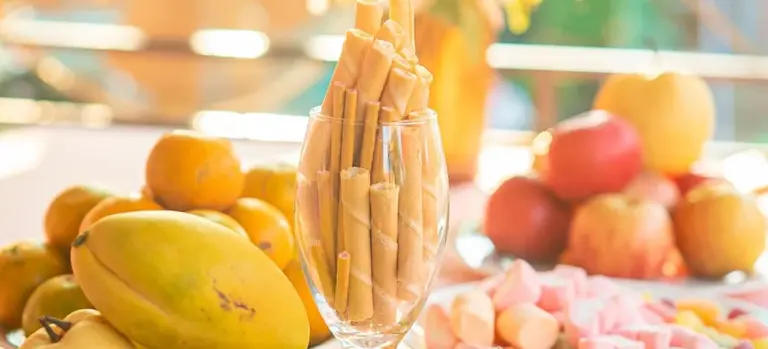 A glass bowl filled with breadsticks sits on a table surrounded by fresh fruit such as mangoes and apples, with other snacks and more fruit in the background.