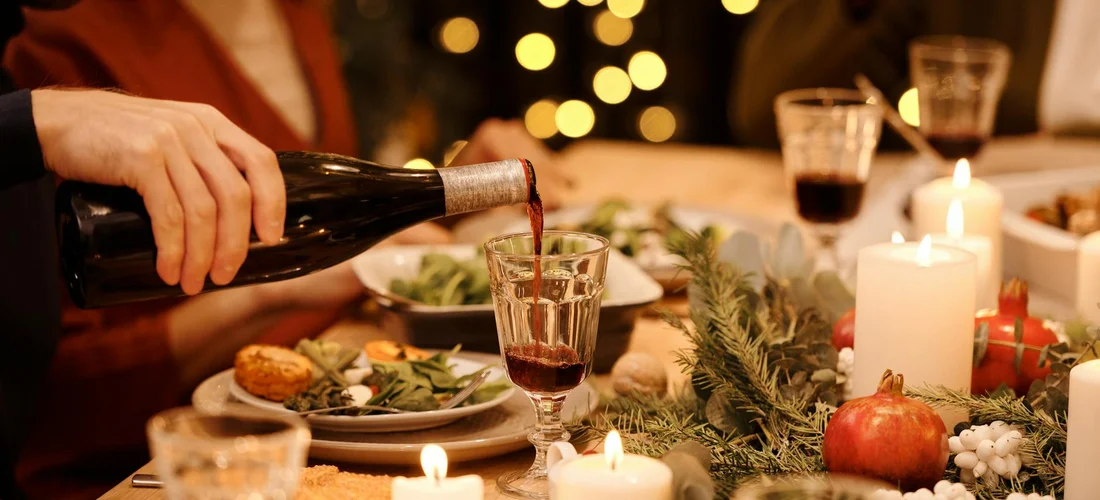 A person pours red wine into a glass at a candlelit dining table with plates, greenery, and candles, suggesting a post-funeral meal.