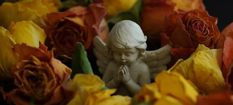 A small angel statue with hands clasped in prayer, surrounded by yellow and orange roses in a funeral or memorial arrangement.