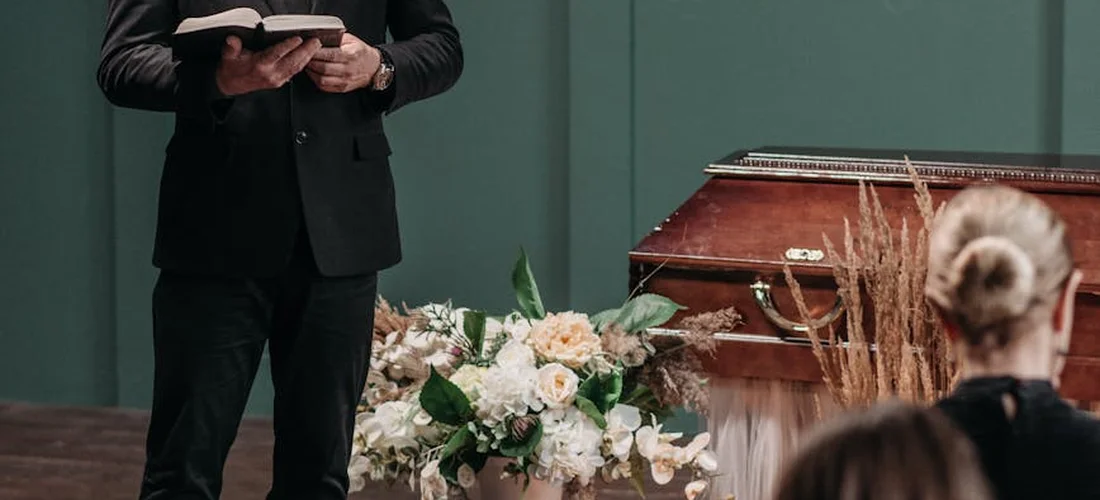 An officiant in a black suit reads from a book at a funeral, with a casket and floral arrangements visible nearby.