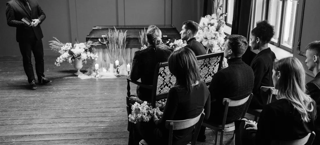 A group of mourners dressed in black sit in a row in a dim room, listening to a speaker at the front of a funeral service.