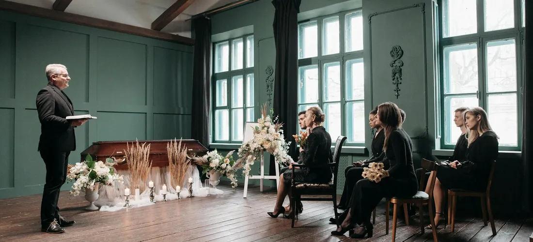 A funeral service scene with a speaker at the front and mourners seated in a small room, dressed in dark clothing, with floral arrangements and candles nearby.