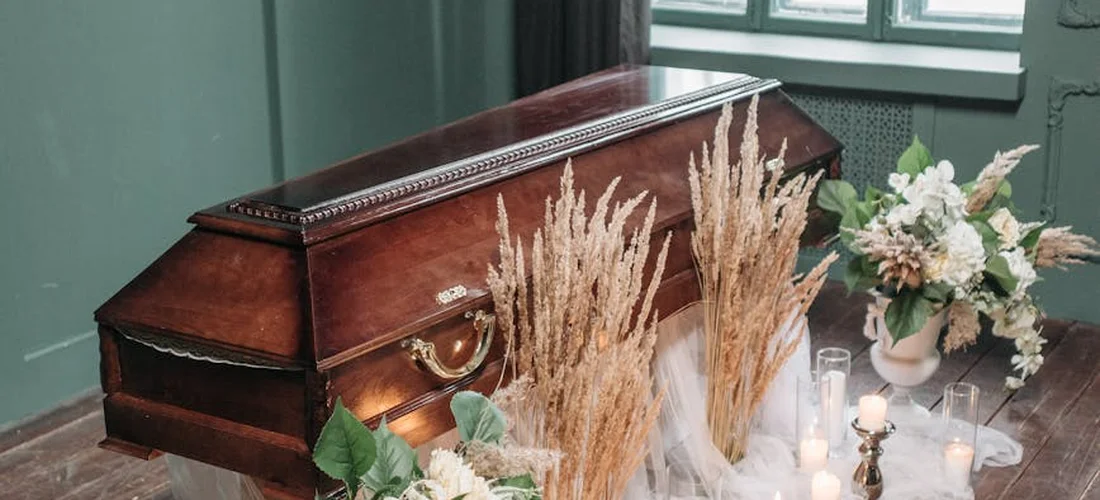 A closed wooden coffin surrounded by dried grasses, candles, and soft floral arrangements in a muted, respectful funeral setting.