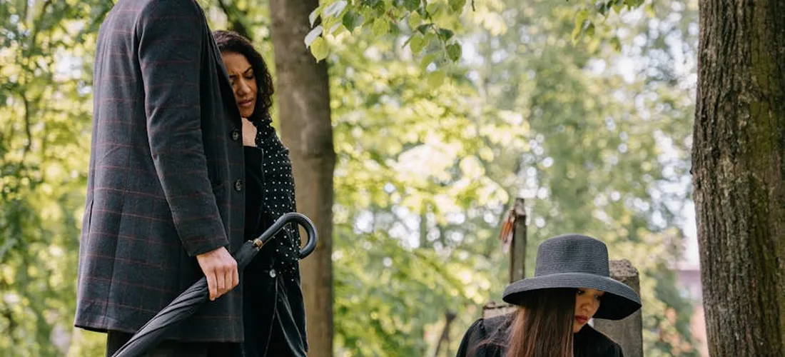 Outdoor cemetery scene with mourners dressed in dark clothing, including a person wearing a wide-brimmed hat.