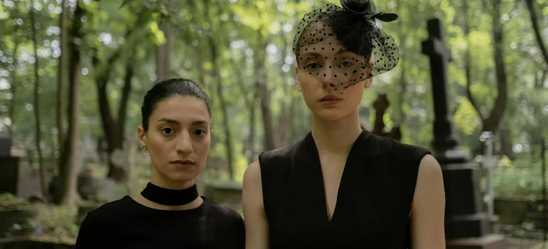 Two women dressed in black at a cemetery visitation; one wearing a sleeveless V-neck dress and the other a black top with a dotted veil, both with solemn expressions.