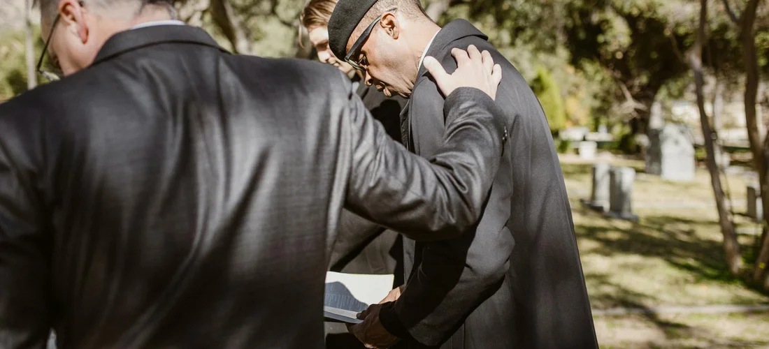 Mourners in dark suits sharing a supportive moment at an outdoor funeral visitation.