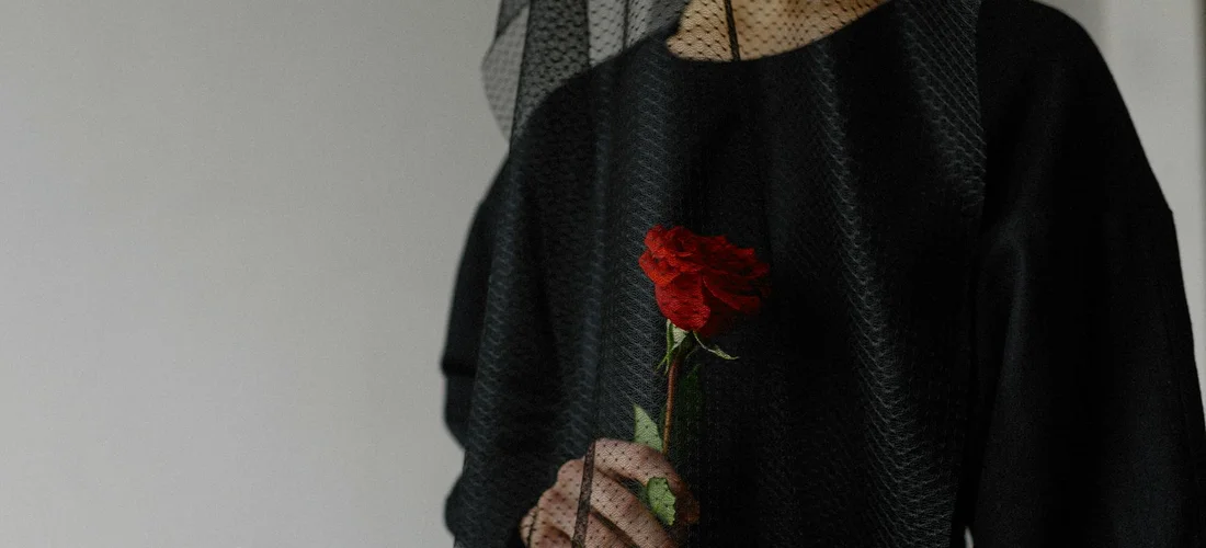 A person wearing black attire and a veil, holding a single red rose.