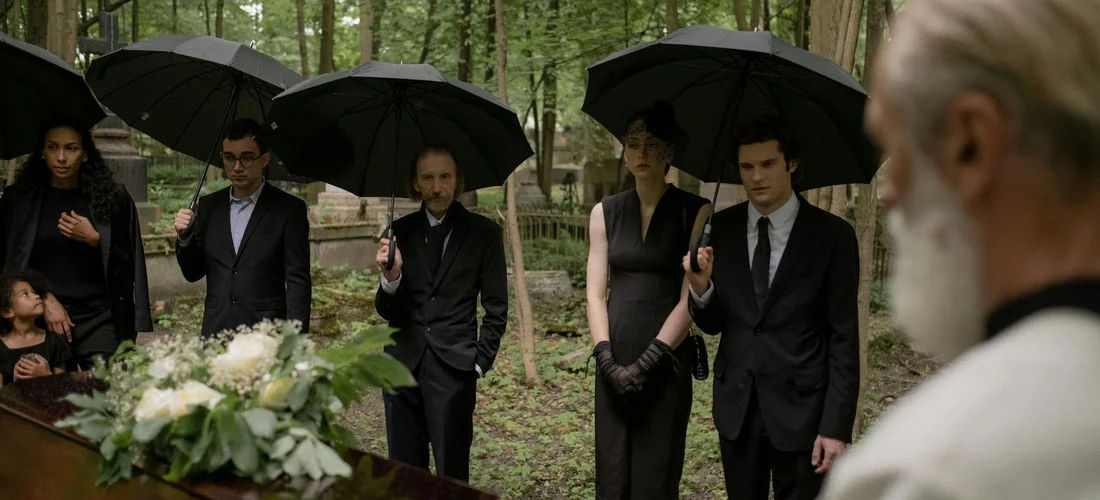 Group of mourners in black attire standing with umbrellas around a casket in a wooded cemetery, with an older officiant on the right.