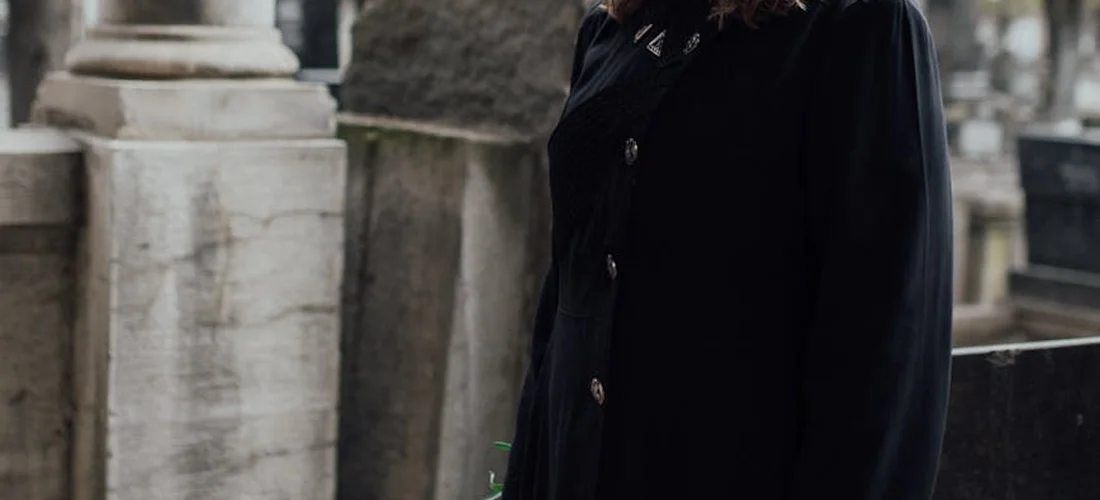 Mourner in a dark coat standing near weathered gravestones in a cemetery, conveying a somber funeral setting.