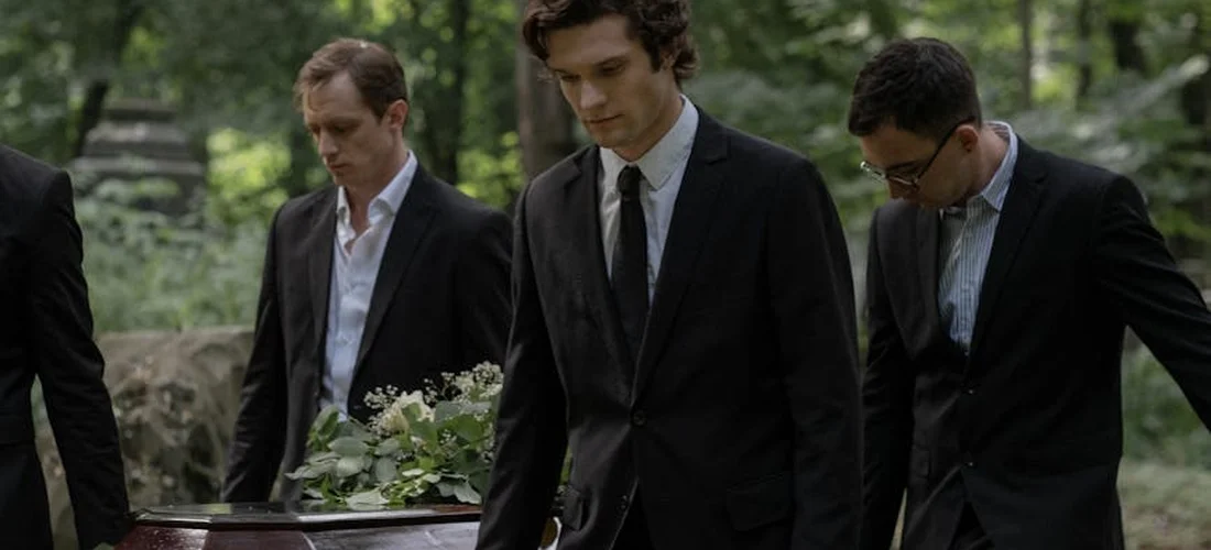 Three men in black suits and ties stand beside a casket adorned with white flowers at a cemetery, capturing the restrained, solemn dress of funeral attire.