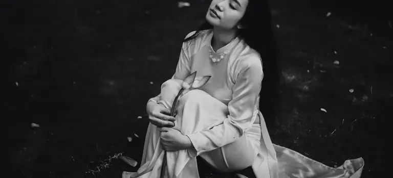 Monochrome image of a woman in a long, modest dress sitting on the ground with a contemplative expression, conveying sobriety and respect.